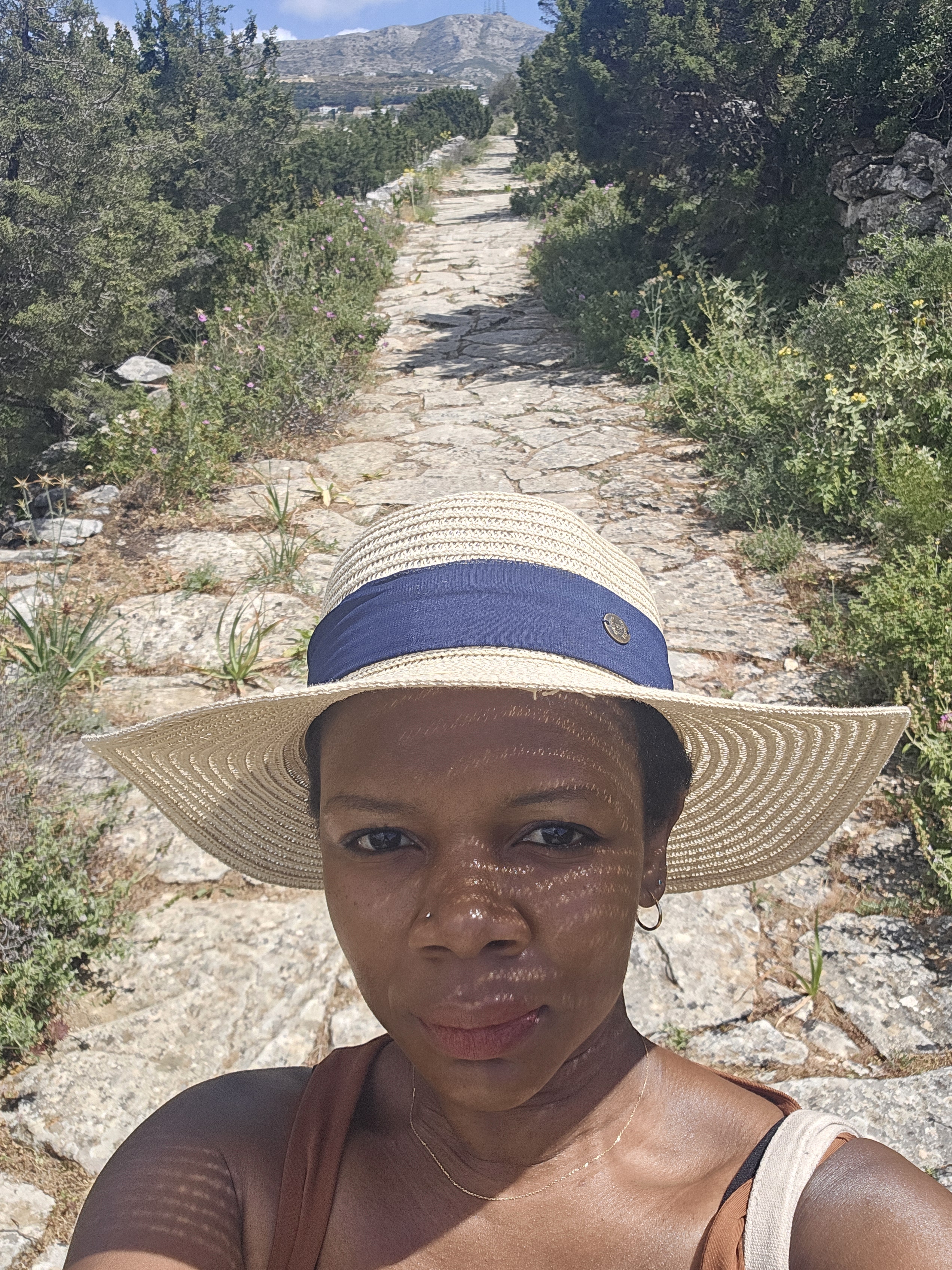 A woman wearing a wide-brimmed straw hat stands on an ancient stone path surrounded by greenery, looking directly at the camera.