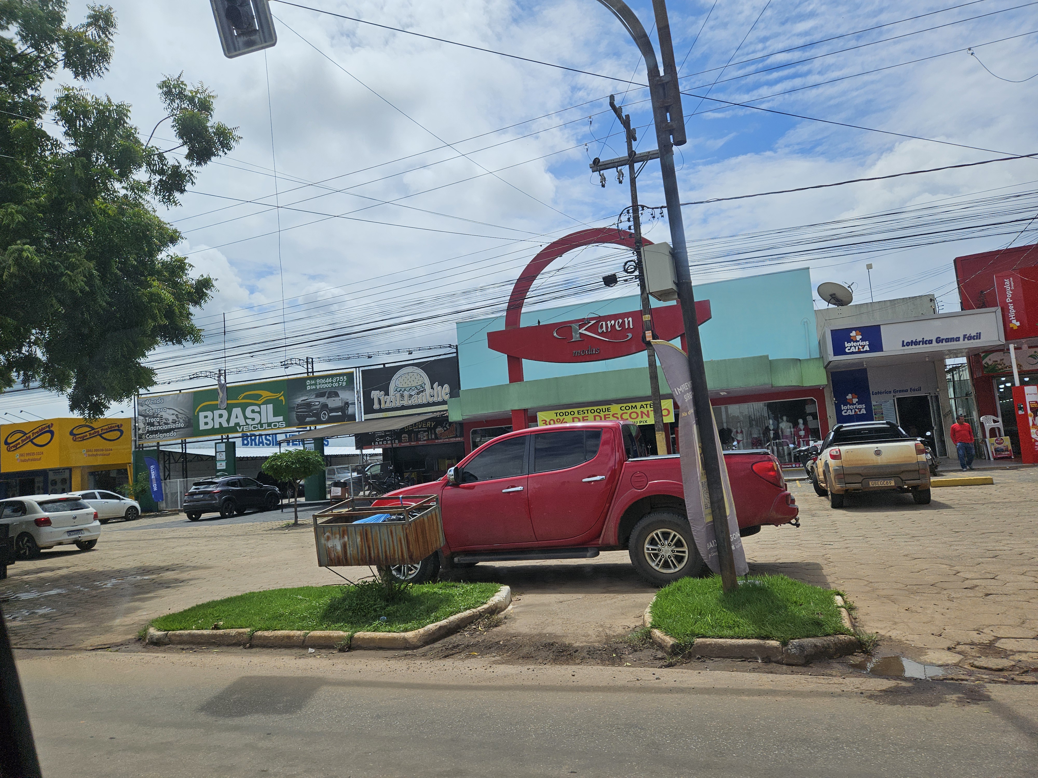 A view of a street scene in Brazil featuring a red pickup truck parked near storefronts, with banners and signs in Portuguese.