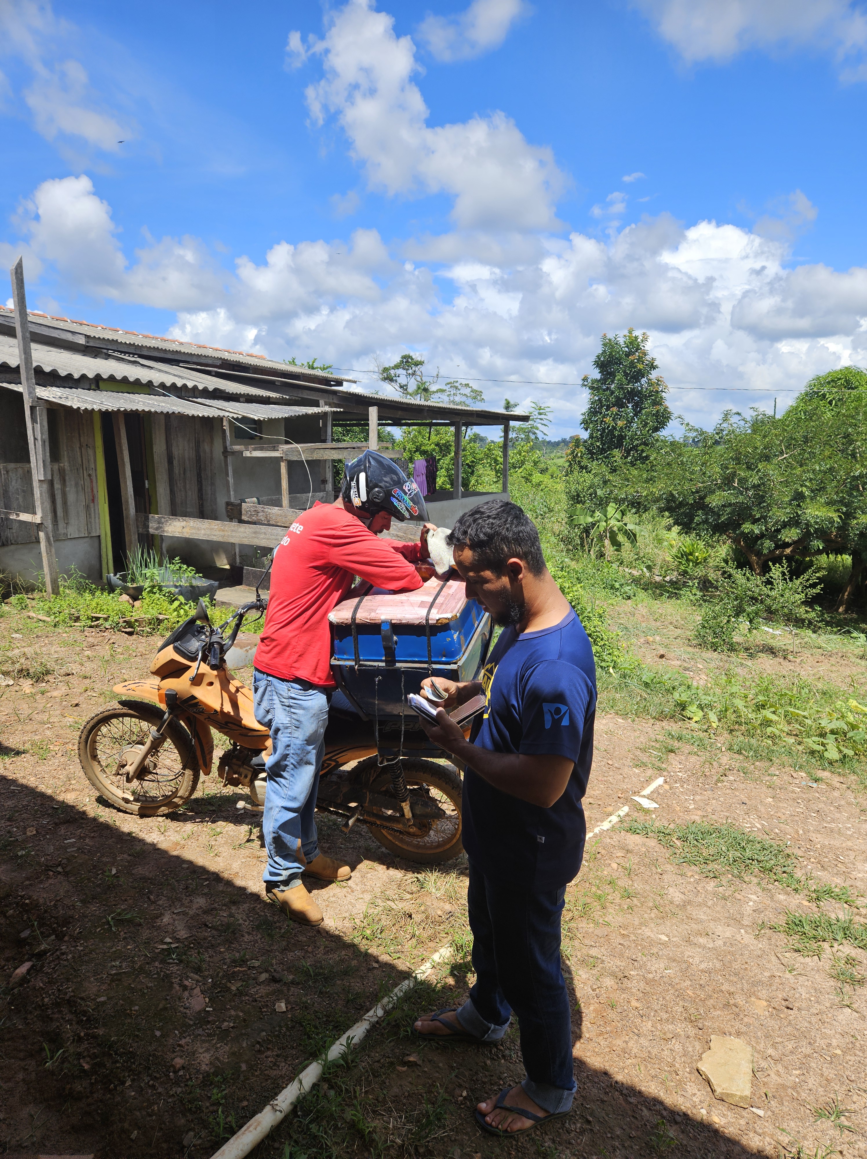 Two men interact outside a rural house in Brazil; one man in a red shirt is preparing to deliver items from a motorbike, while the other checks his phone.