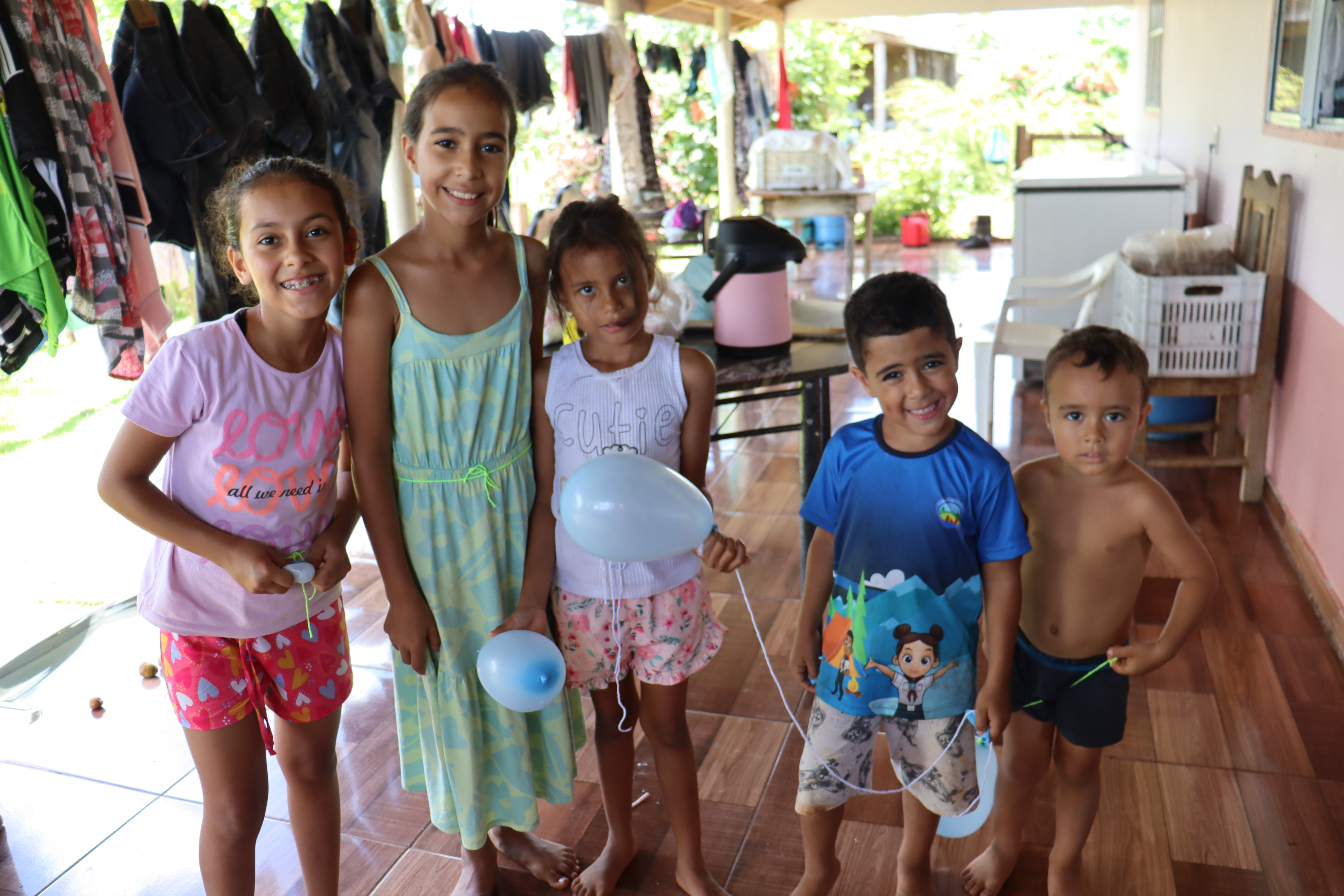 A group of five children smiling and posing for a photo indoors, surrounded by clothing hung out to dry in the background.