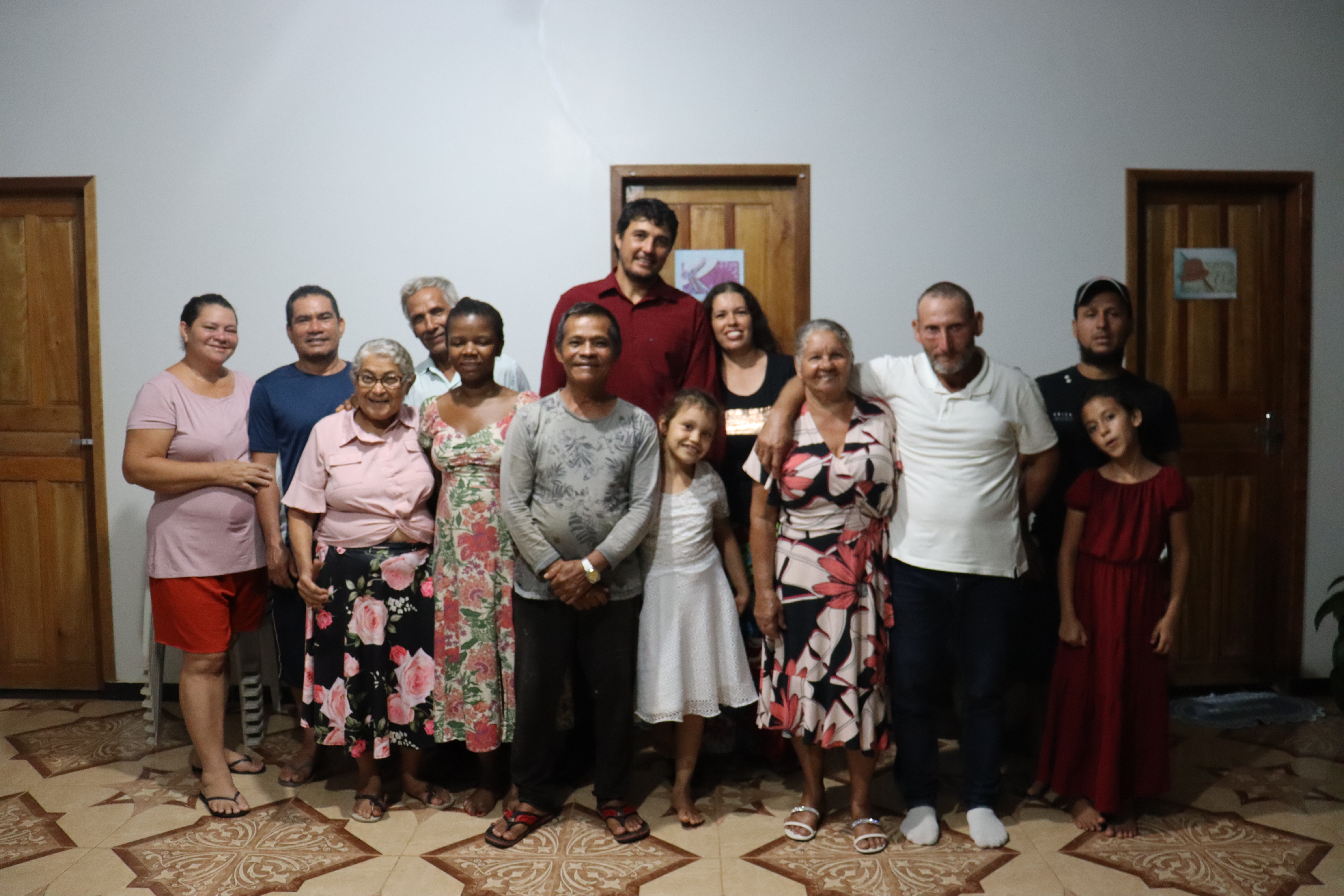 A group of people posing together in a room, smiling and standing closely. The background features wooden doors and a patterned floor.